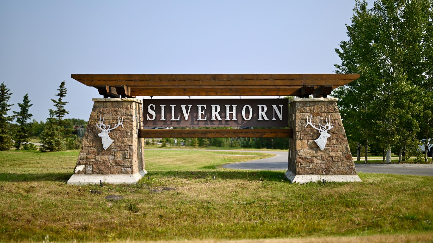 The Silverhorn entrance sign: a heavy timber beam on two stone piers, each carrying a stag in relief, with the community road running behind it.